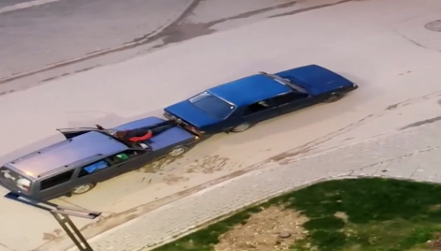 Otomobilin kaputuna oturdu, ayaklarıyla öndeki aracı itti